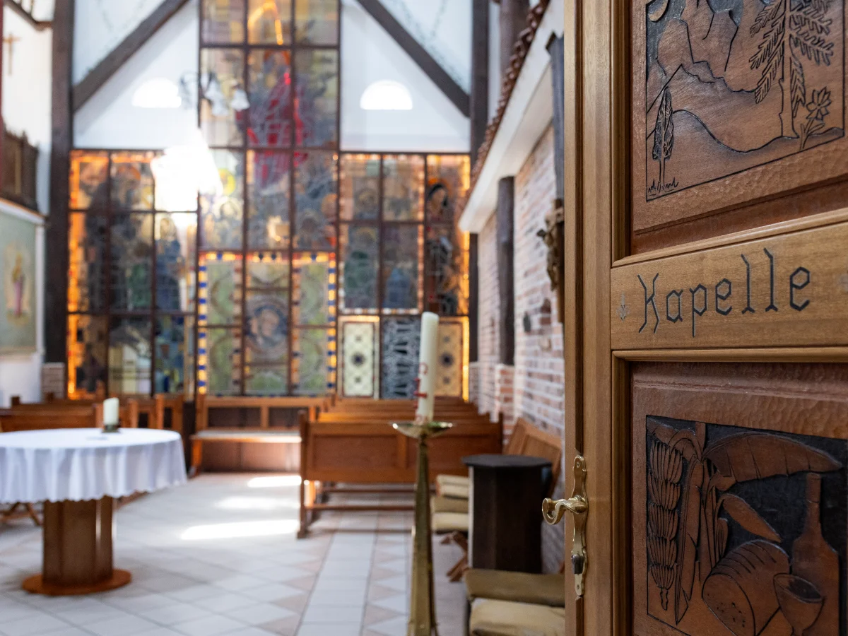 Blick in die Kapelle des St. Josef Stifts, mit einem Holztisch und Bänken vor farbigen Glasfenstern.