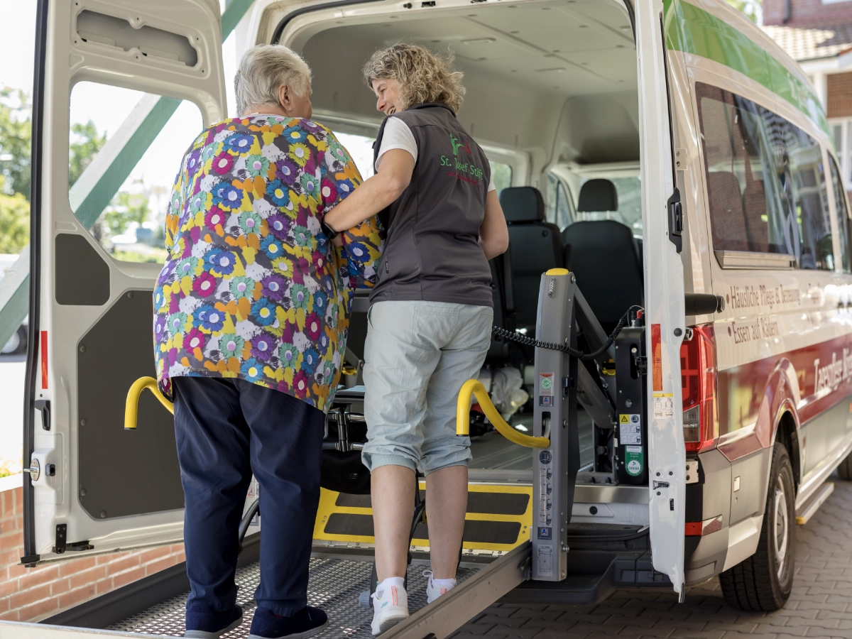 Eine Frau vom Fahrdienst hilft der Klientin über die Einstiegshilfe ins Auto.