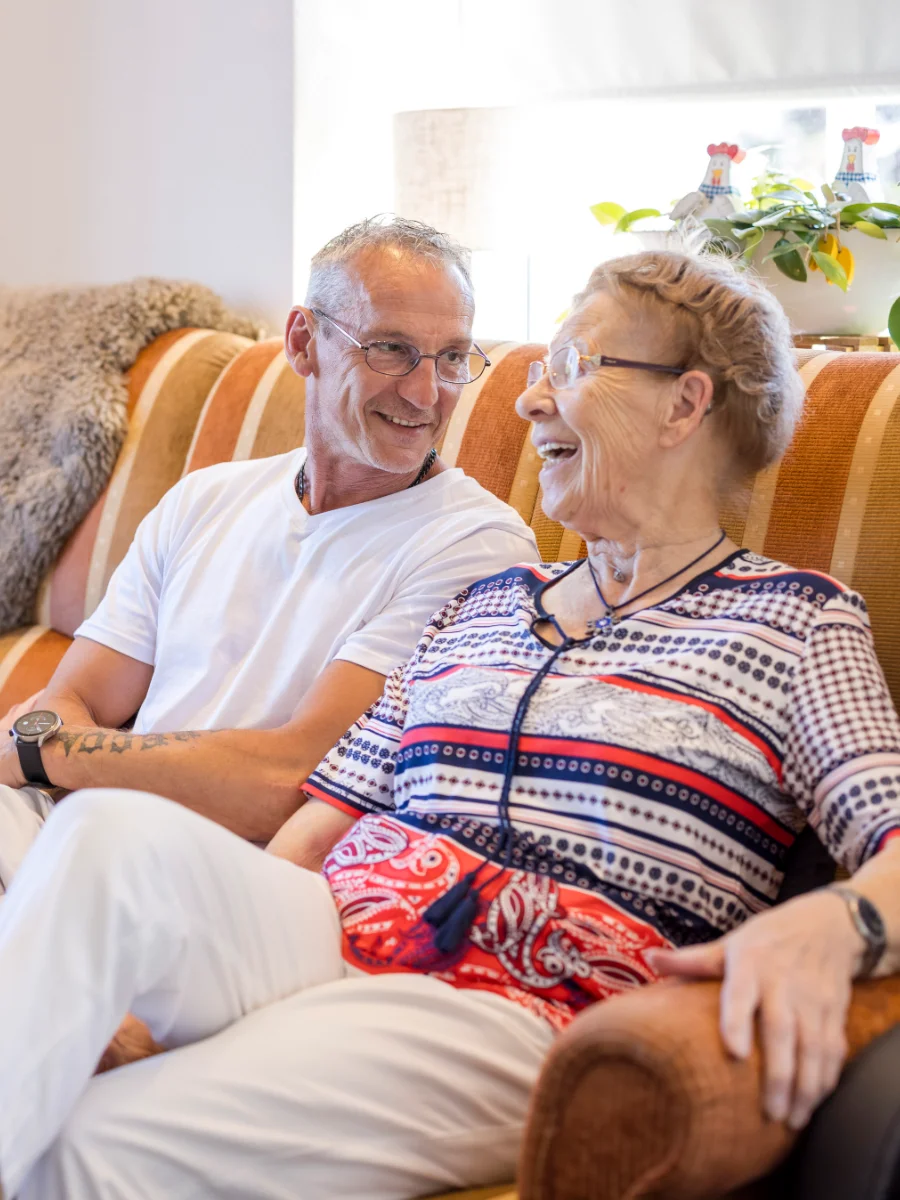 Ein Pfleger und eine ältere Dame sitzen lachend auf einem Sofa.