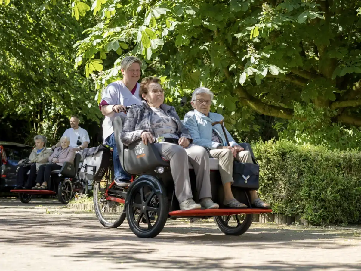 Pflegerin von wi kümmert us fährt zwei Seniorinnen in einem roten Fahrradtaxi durch einen sonnigen Park
