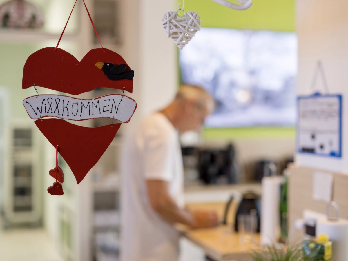 Ein rotes Schild in Herzform mit einem schwarzen Vogel darauf. Auf dem Schild steht in Handschrift 'Willkommen'. Im Hintergrund sieht man verschwommen einen Pfleger.